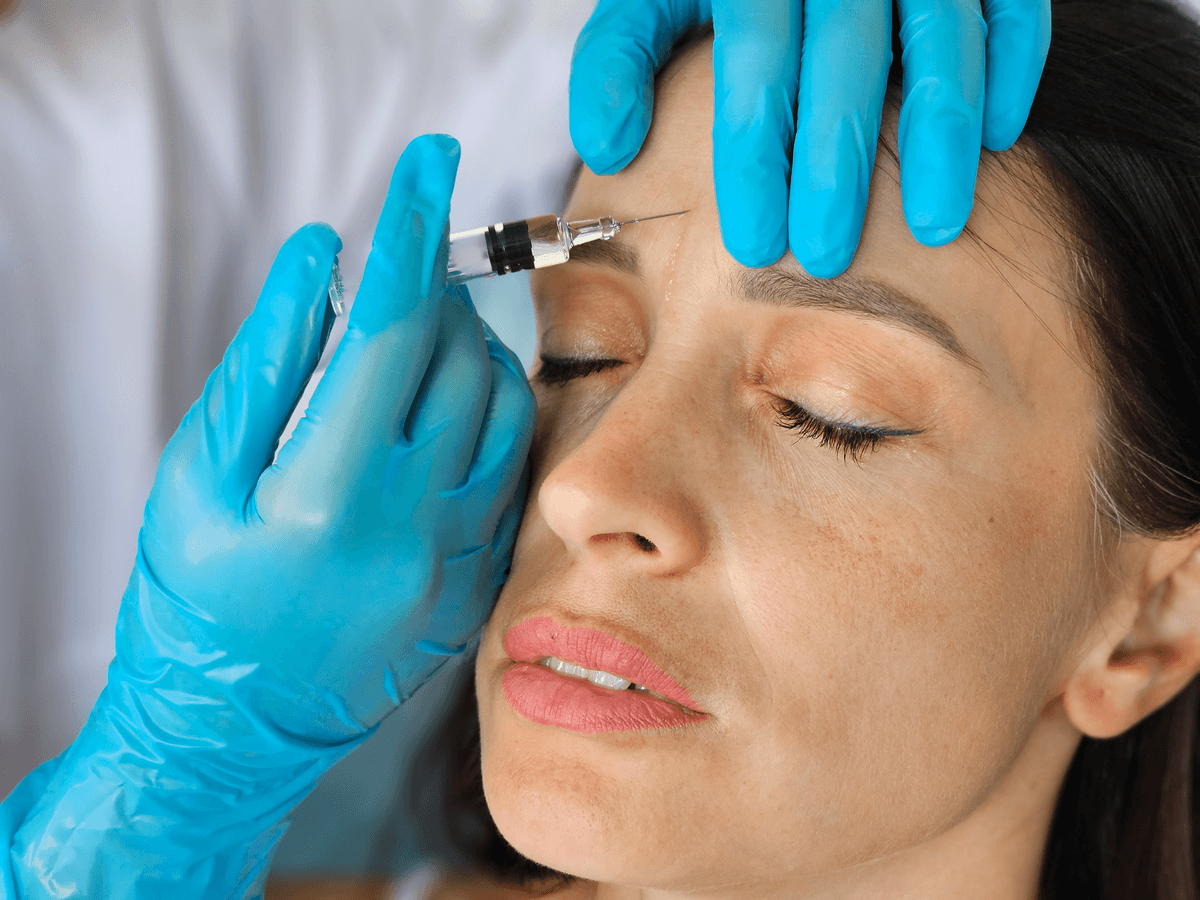 Lékař v rukavicích aplikuje botulotoxin injekční stříkačkou nad obočí